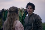 "A young man in old-fashioned clothing stands outside facing a woman with long hair, both appearing somber. Behind them are green leaves hanging, possibly on a rural farm, under a cloudy sky during the day."