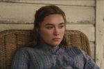 "A young woman in a blue patterned dress sits pensively in a wicker chair against a worn wooden wall, appearing thoughtful and somber in a rustic, vintage setting."