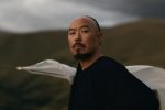"A man with a mustache and goatee stands outdoors against rolling hills and cloudy sky, dressed in dark clothing with a white cloth behind him, looking toward the camera in a natural, rugged landscape."