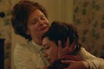 "Two women in vintage clothing share an emotional embrace in a warmly lit, floral-wallpapered room, suggesting comfort and support in a historical or period setting."