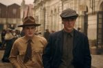"Two young men in vintage clothing and hats walk on an old city street with historical buildings and people in period attire in the background, evoking an early 20th-century urban setting."