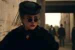 "Woman in vintage black attire with round sunglasses, a veiled hat, and gold jewelry stands near a stone wall in an urban setting, with blurred people and old-style architecture in the background."